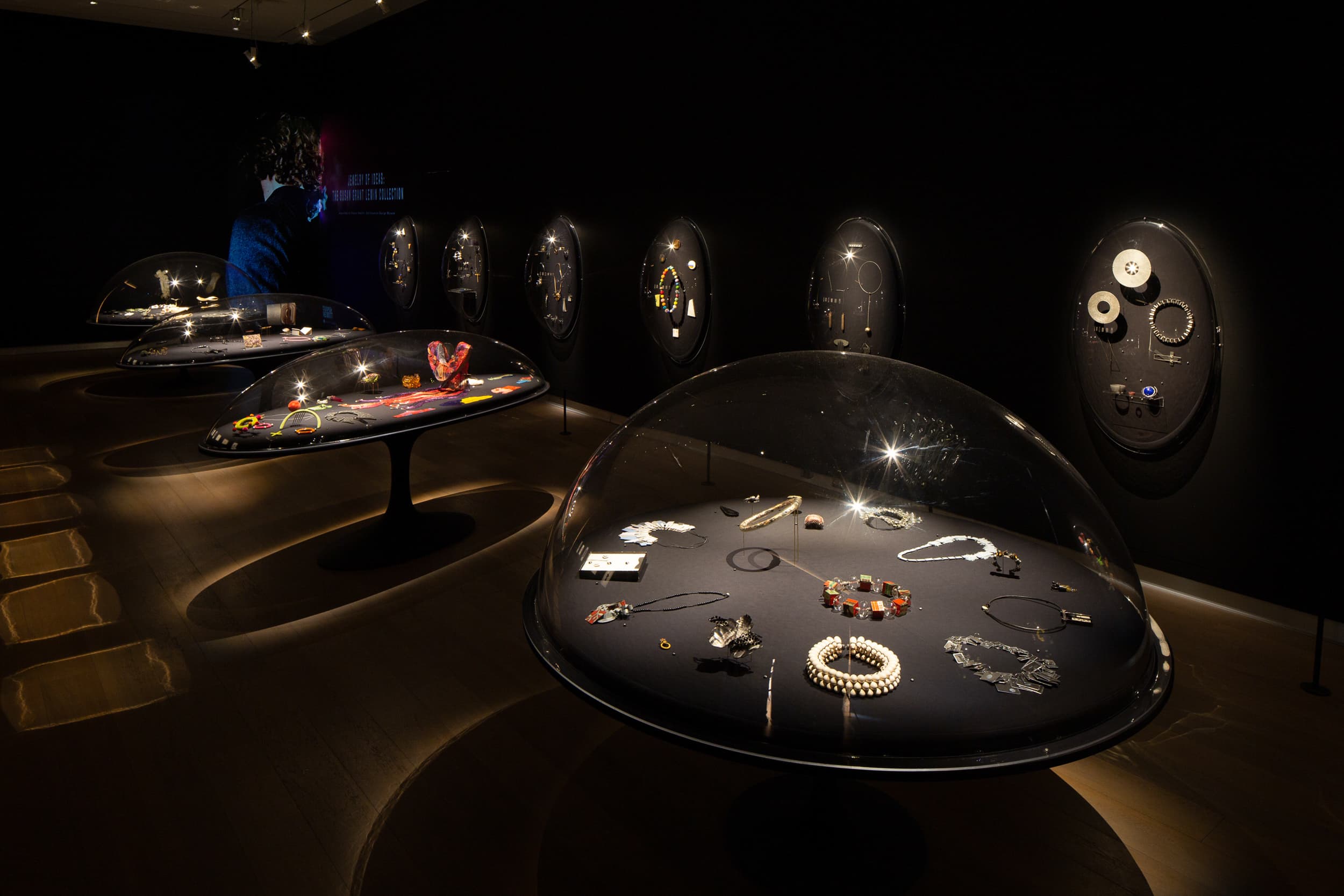 Dark jewelry exhibition with accessories displayed under clear domes and on circular wall panels — before retouching